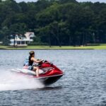Person riding a red jet ski across a lake, with water spraying behind, trees, and a house visible in the background. Person riding a red jet ski across a lake, with water spraying behind, trees, and a house visible in the background.