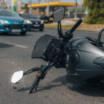 A fallen motorcycle lies on its side on a busy road, with cars and a gas station in the background, conveying a scene of a recent accident.
